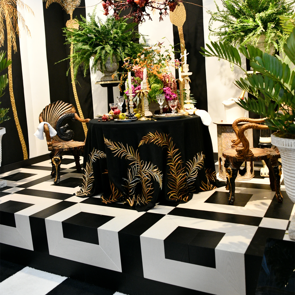 A styled interior display with a black-and-white geometric platform, a black draped table with gold palm-leaf embroidery, ornate chairs, potted plants, and a large multicolored floral arrangement in the center.